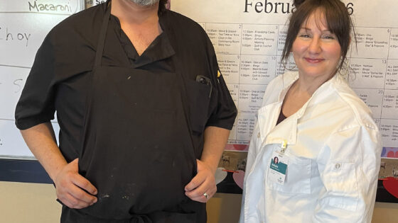 A man, wearing all black and an apron, and a woman, wearing chef's whites, stand side by side smiling at the camera next to a white board listing the day's meal specials.