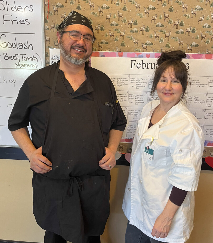 A man, wearing all black and an apron, and a woman, wearing chef's whites, stand side by side smiling at the camera next to a white board listing the day's meal specials.