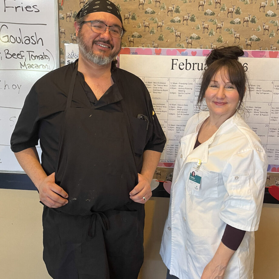 A man, wearing all black and an apron, and a woman, wearing chef's whites, stand side by side smiling at the camera next to a white board listing the day's meal specials.