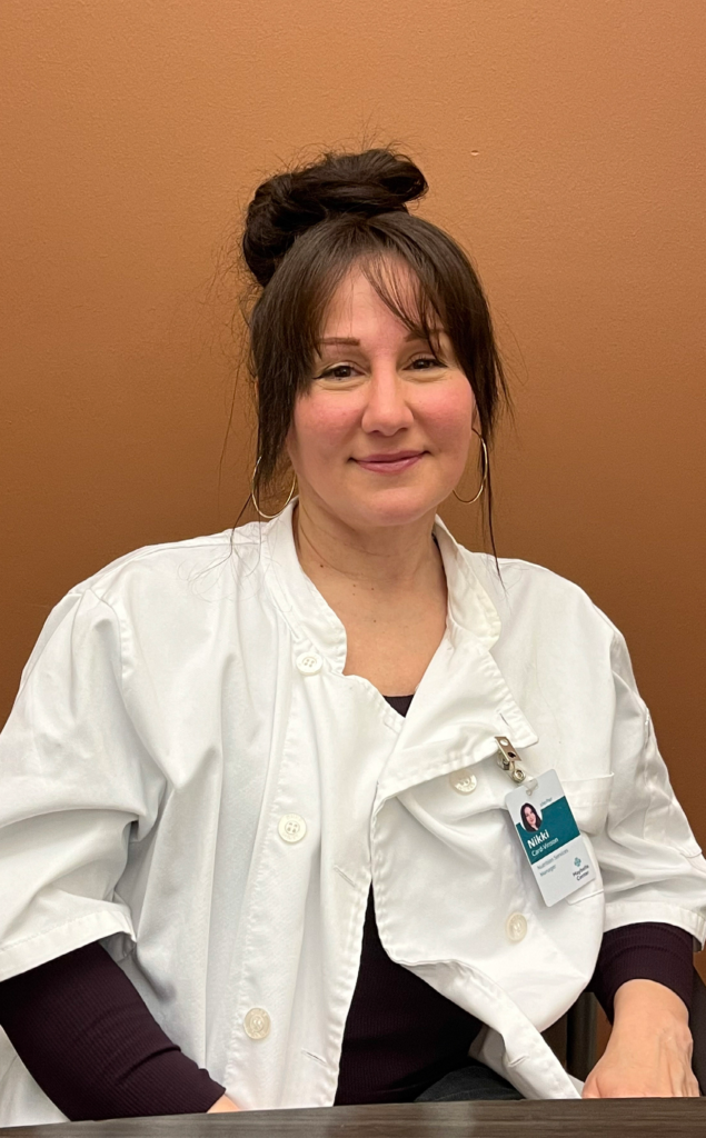 Seated white woman with dark hair up in a bun, softly smiling directly at the camera, wearing chef's whites and a clipped on name badge