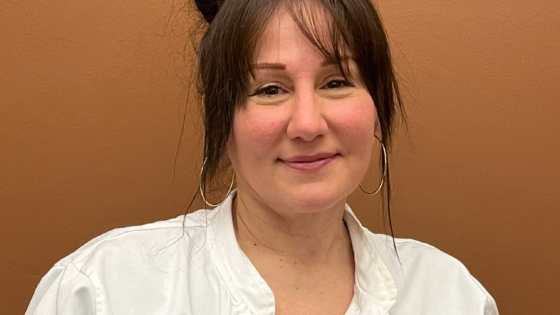 Seated white woman with dark hair up in a bun, softly smiling directly at the camera, wearing chef's whites and a clipped on name badge