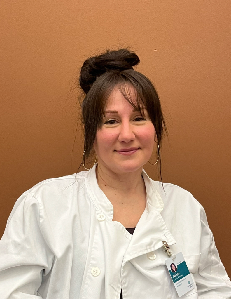 Seated white woman with dark hair up in a bun, softly smiling directly at the camera, wearing chef's whites and a clipped on name badge