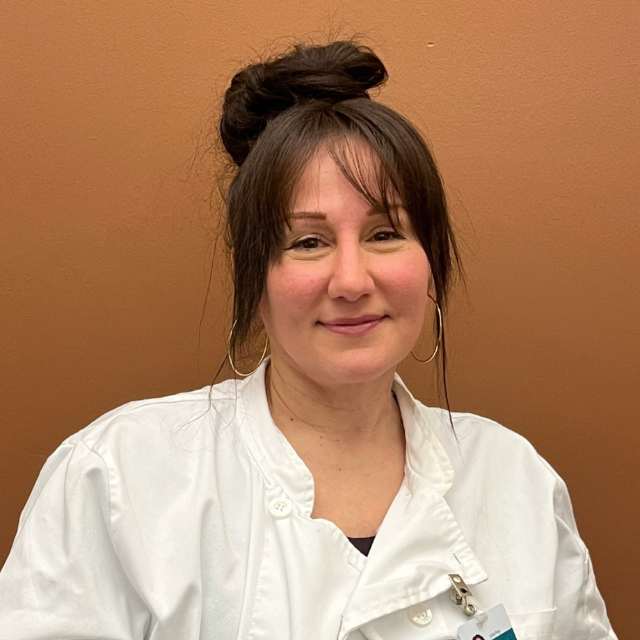 Seated white woman with dark hair up in a bun, softly smiling directly at the camera, wearing chef's whites and a clipped on name badge