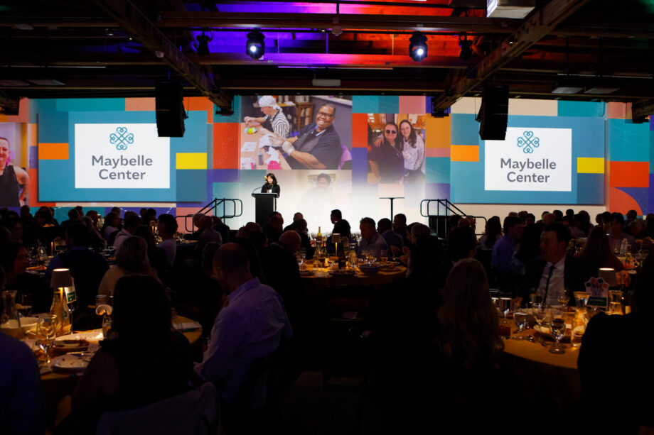 Room full of multiple tables of people seated around them, looking towards the wall where a photo display of images is projected and two screens on either side of a podium show the Maybelle Center logo.