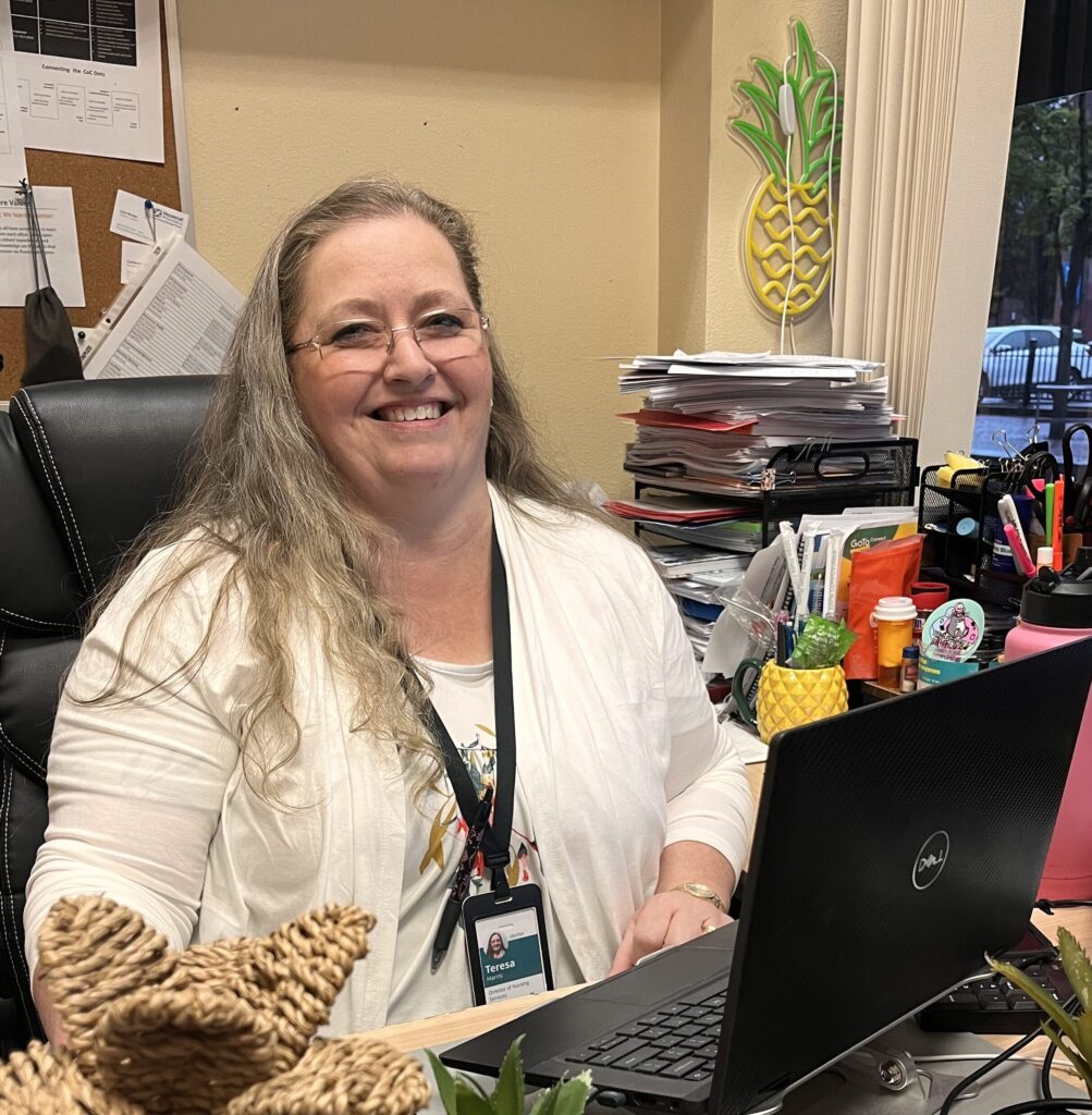 Middle aged white woman with long hair sits at a desk in front of an open laptop, smiling at the camera