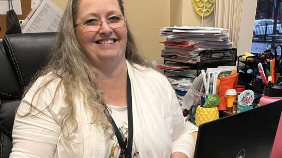 Middle aged white woman with long hair sits at a desk in front of an open laptop, smiling at the camera