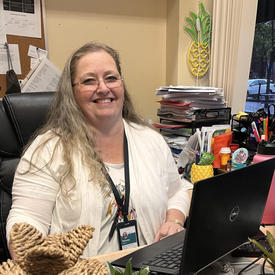 Middle aged white woman with long hair sits at a desk in front of an open laptop, smiling at the camera