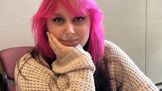 Young woman with pink and dark hair and cat-eye eye make up, sitting at a table, with her face in one hand, looking at the camera with a soft smile