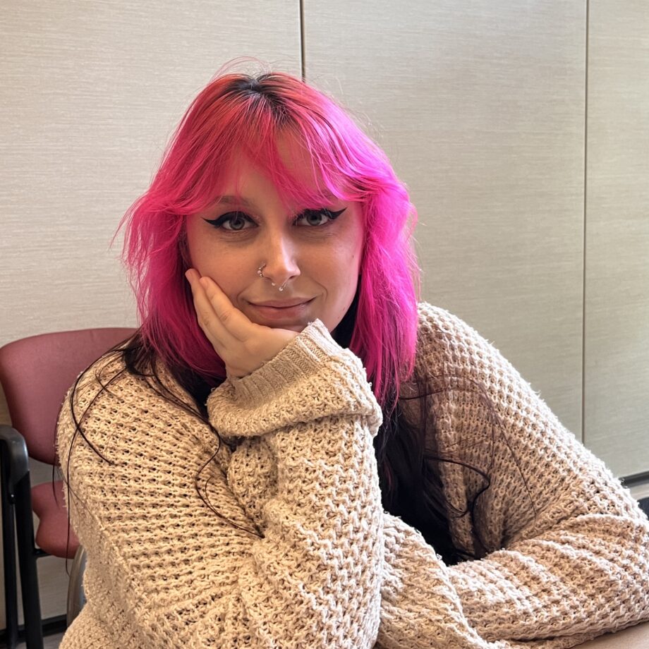 Young woman with pink and dark hair and cat-eye eye make up, sitting at a table, with her face in one hand, looking at the camera with a soft smile