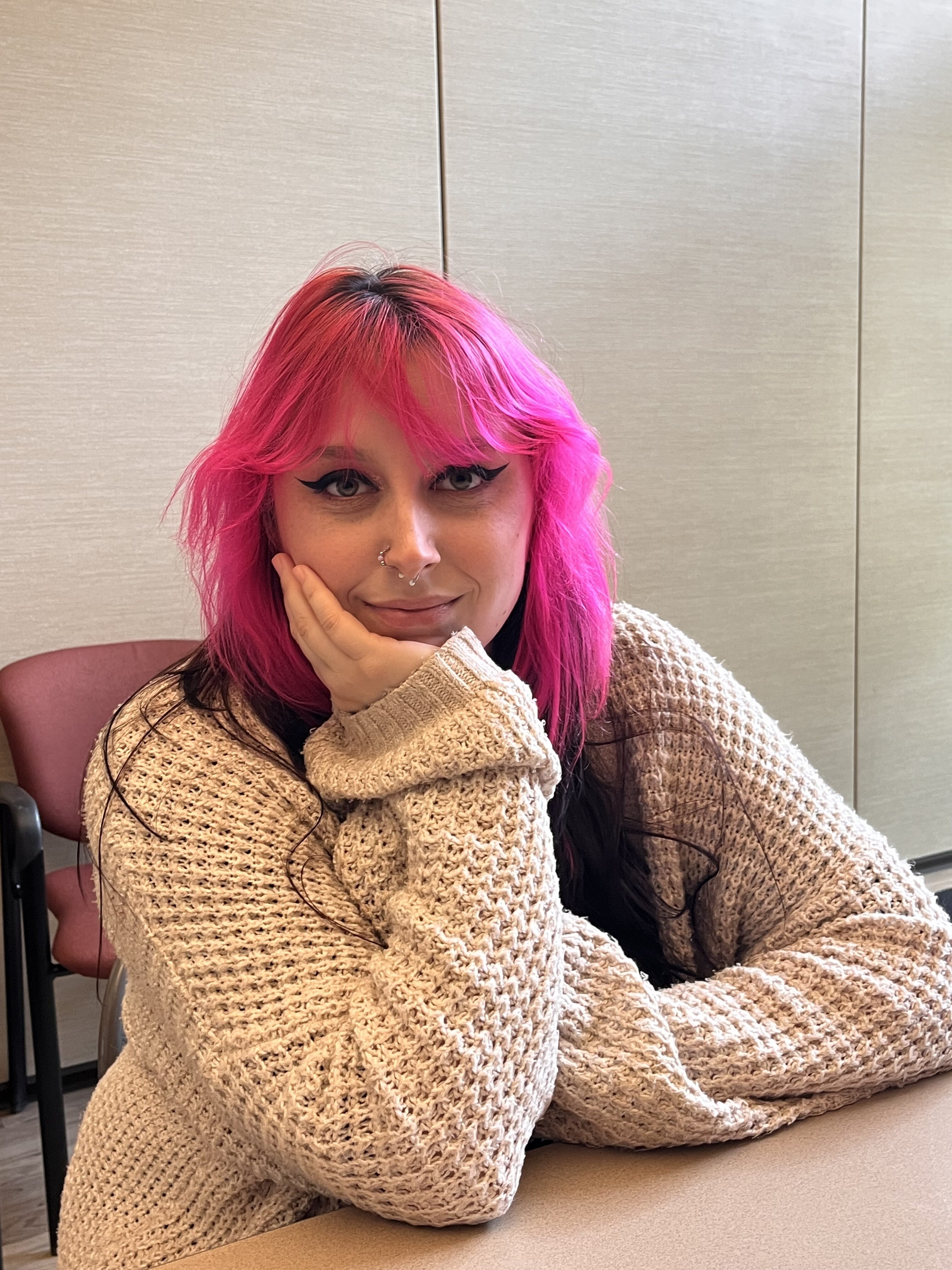 Young woman with pink and dark hair and cat-eye eye make up, sitting at a table, with her face in one hand, looking at the camera with a soft smile