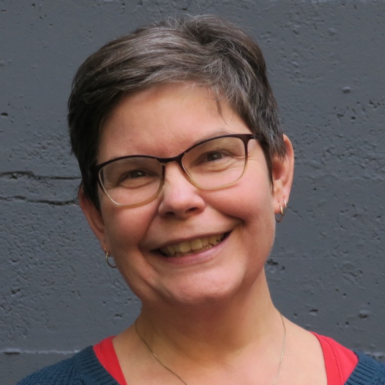 White woman with close-cropped salt & pepper hair, smiles directly into the camera wearing rectangular-rimmed glasses