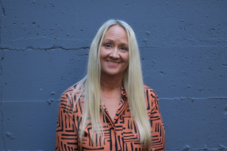 White woman in a professional blouse with long bleached blond hair standing in front of a painted cement wall, smiling directly at the camera