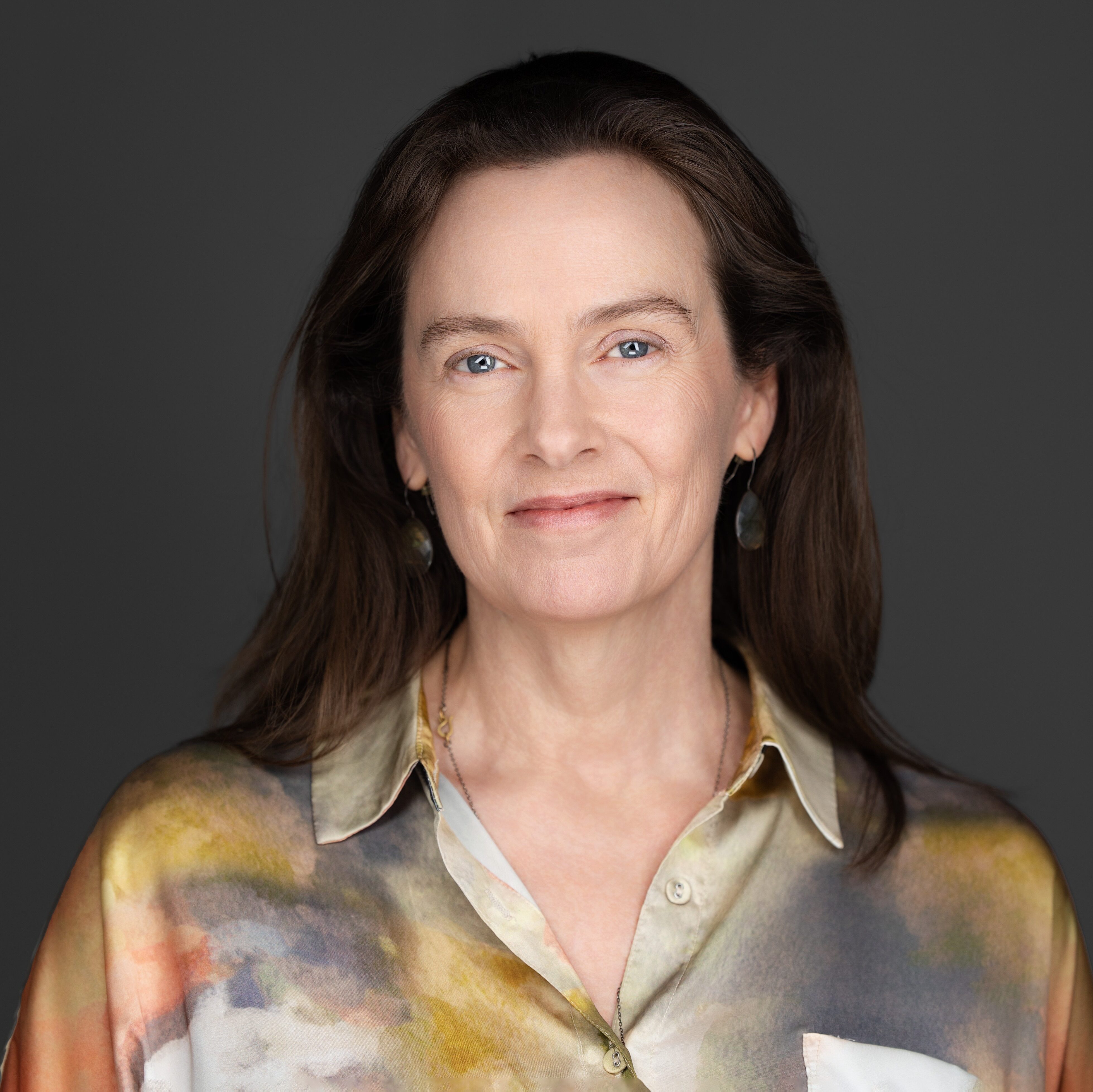 White woman with shoulder-length brown hair smiling with closed mouth directly at camera, wearing silky blouse
