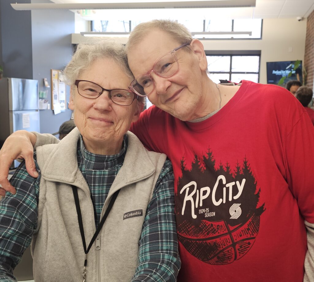 Older white man stands with arm around shoulders of older white woman, both smile directly to the camera and have the sides of their foreheads pressed together