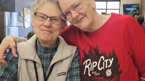 Older white man stands with arm around shoulders of older white woman, both smile directly to the camera and have the sides of their foreheads pressed together