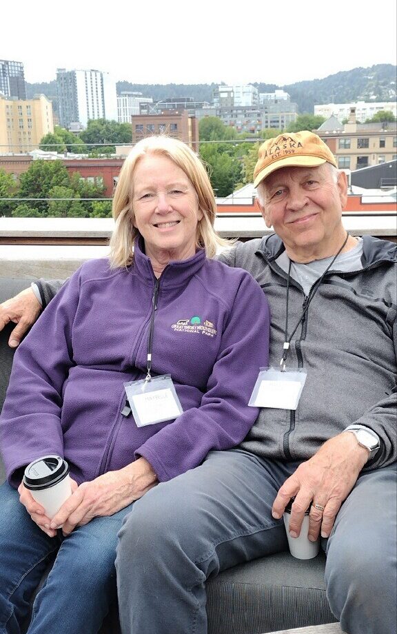 Older white woman and white man sit side by side leaning into each other, wearing name badges around their neck, sweaters, and jeans, while smiling at the camera