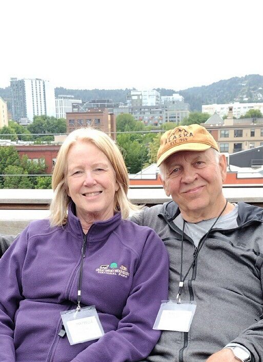Older white woman and white man sit side by side leaning into each other, wearing name badges around their neck, sweaters, and jeans, while smiling at the camera