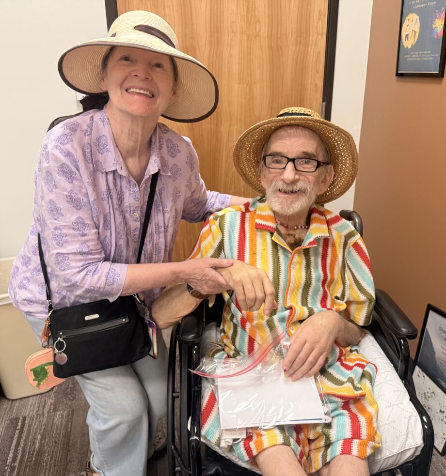 White woman smiles at camera, crouching next to and gently holding the hand of a white, older man who is seated in a wheelchair and wearing colorful striped ensemble, also smiles into the camera.