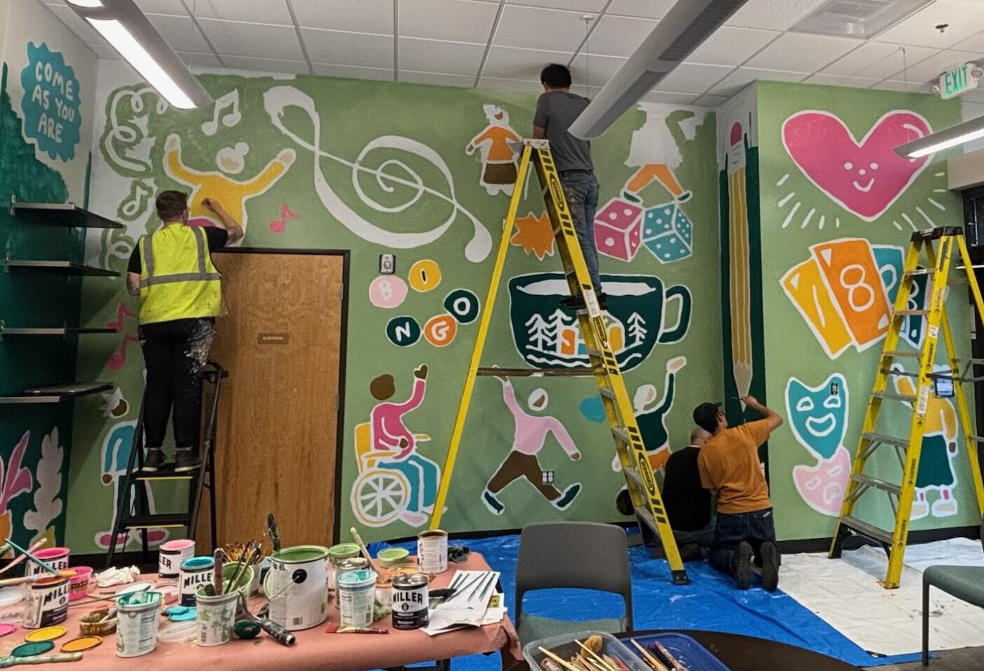 Adults stand, kneel, and balance on a ladder, all painting collaborative on a bright mural inside Maybelle Center's Community Room