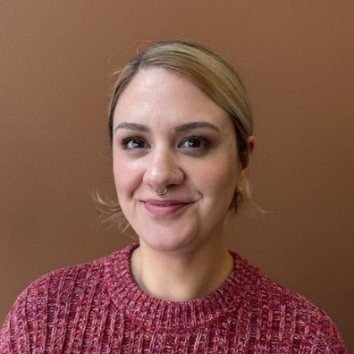 Headshot of a young white woman smiling directly into the camera with light hair pulled back behind her.